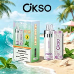 Un kit da vaporizzatore OKSO Miami Mint in scatola su una spiaggia sabbiosa con una bottiglia di menta e un dispositivo con motivi viola pastello, palme e oceano blu sullo sfondo.