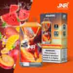 Un'immagine promozionale che mostra un prodotto da vaporizzare etichettato "JNR" al sapore di anguria, mango e pesca. Include un disegno colorato di un uccello alato sulla confezione del prodotto, circondato da immagini di frutta fresca e schizzi di liquidi, con un'etichetta di avvertenza sulla dipendenza da nicotina sulla scatola.