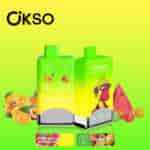Two brightly colored OKSO vape devices with green-yellow gradient packaging, one featuring a cartoon monkey and the other showing icons for mixed fruit and watermelon bubble gum flavors, surrounded by images of assorted fruits and candies, with product flavor labels and branding on a yellow-green background.