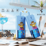 Two blue BANG DE vape devices with a cartoon monkey wearing a crown, posed on a table with a cocktail, lemon slices, and blurred beachside background, highlighting '3 IN 1', '80,000 PUFFS', and product features.
