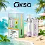 A boxed OKSO Miami Mint vape kit on a sandy beach with a minty bottle and a pastel purple patterned device, palm trees and blue ocean in the background.