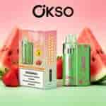 Strawberry watermelon OKSO vaping device with green patterned panel beside a matching box, set against pink dessert-themed background with watermelon slices and strawberries.