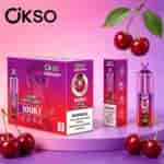Sfondo rosa-viola brillante con diverse scatole viola OKSO Tornado e ciliegie mature appese agli steli, ciliegie sparse sulla superficie.